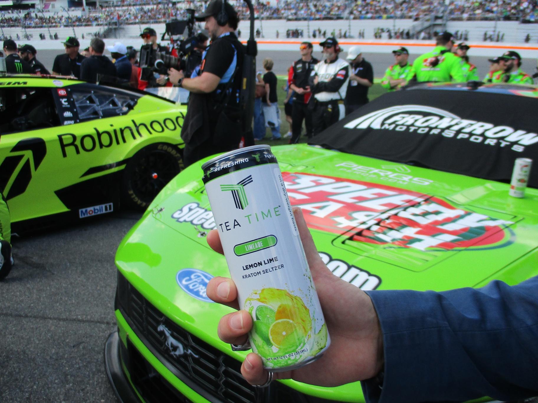 A Tea Time Limeade can held in front of a green race car at the track.